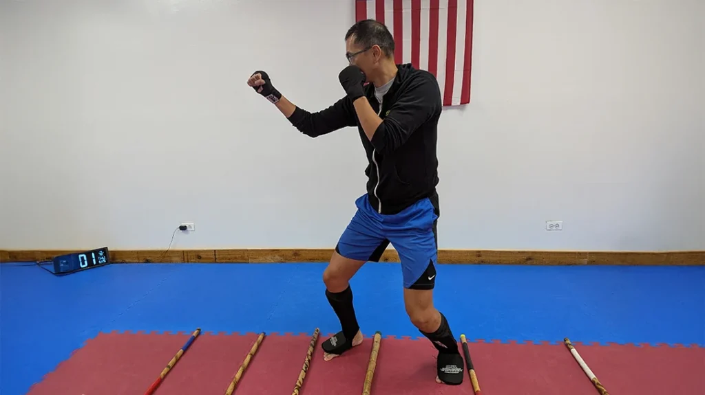 Adam aligns several short sticks to create a makeshift agility ladder. It is even more challenging to train with because he must ensure that he does not touch the sticks to prevent them from moving. He begins in a basic fighting stance. This can be done by drilling forward and backward.